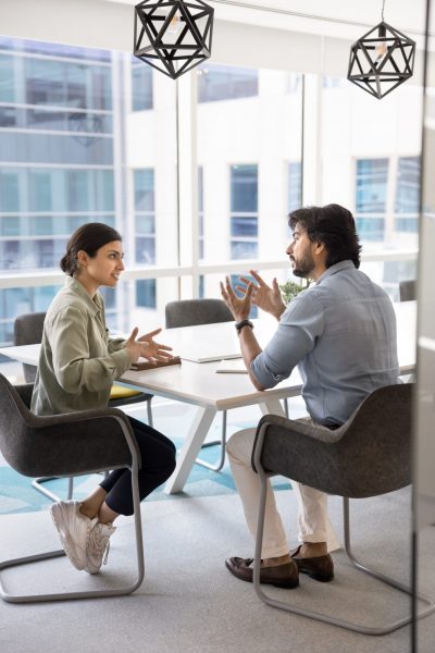 Two young Indian male and female business partners engaged in conversation sitting at table, talking in meeting room, discussing work project, cooperation, partnership. Vertical full body shot