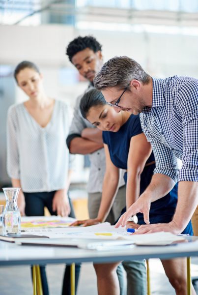 Male and female managers planning at a table with younger staff members looking on.